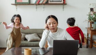 Mom struggling with finding balance in life sitting at a computer appearing stressed as 2 kids run around in the background behind her