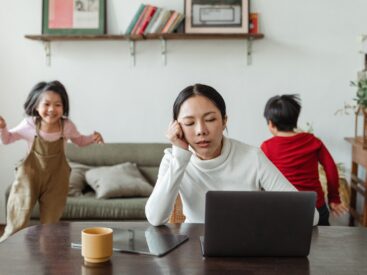 Mom struggling with finding balance in life sitting at a computer appearing stressed as 2 kids run around in the background behind her