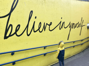 Little boy in a yellow and navy blue coat looking up at a yellow wall with the words believe in yourself painted on it to symbolize need to master your mindset