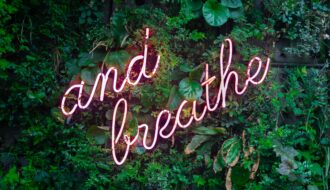 pink neon sign that says Just breathe and hanging on a green planted wall for a blog article about how to show up for yourself