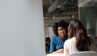 A black woman and a white woman sitting at a table across from each other for a blog article on how to assert yourself