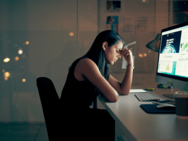 Woman sitting bent over at computer with hand to her temple for blog article about mental exhaustion