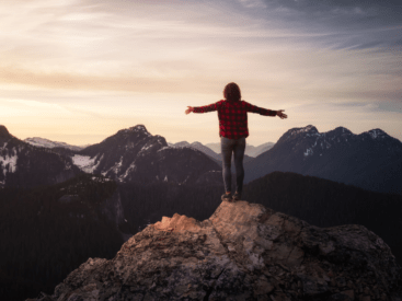 Picture of someone from behind, on a mountain top surrounded by trees with their arms out to their sides, for blog article about transformational coaching