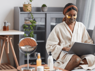 Women sitting on the couch in a rode on her computer with an eye mask on and cosmetics on the table beside her for a blog article about prioritizing yourself