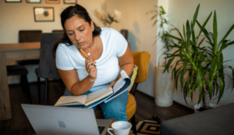 A woman sitting in a chair looking at a laptop screen with a book and notepad on her lap and a pen in her hand for a blog article on how to invest in yourself.